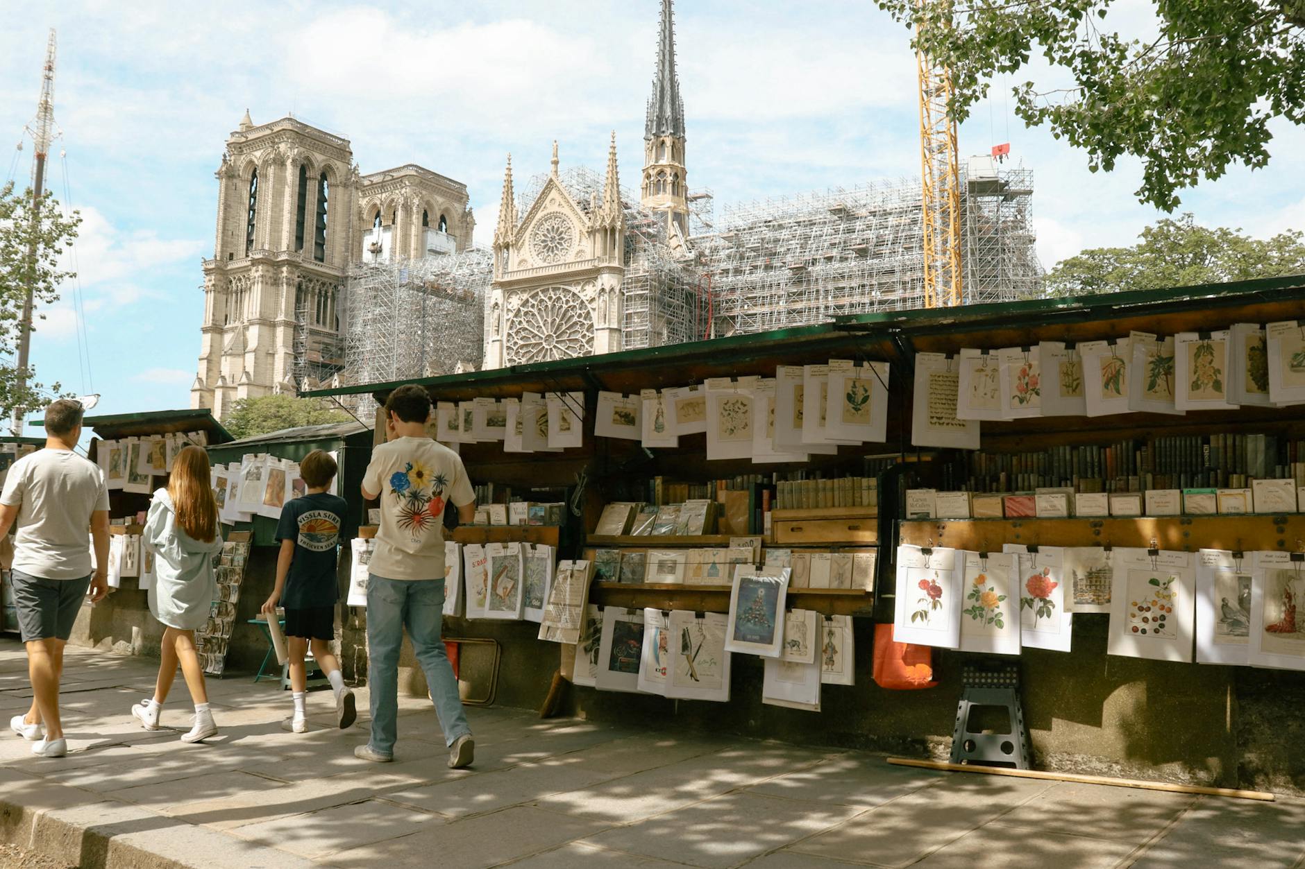 Tourists explore art and book stalls beside Notre-Dame Cathedral in Paris on a sunny day.