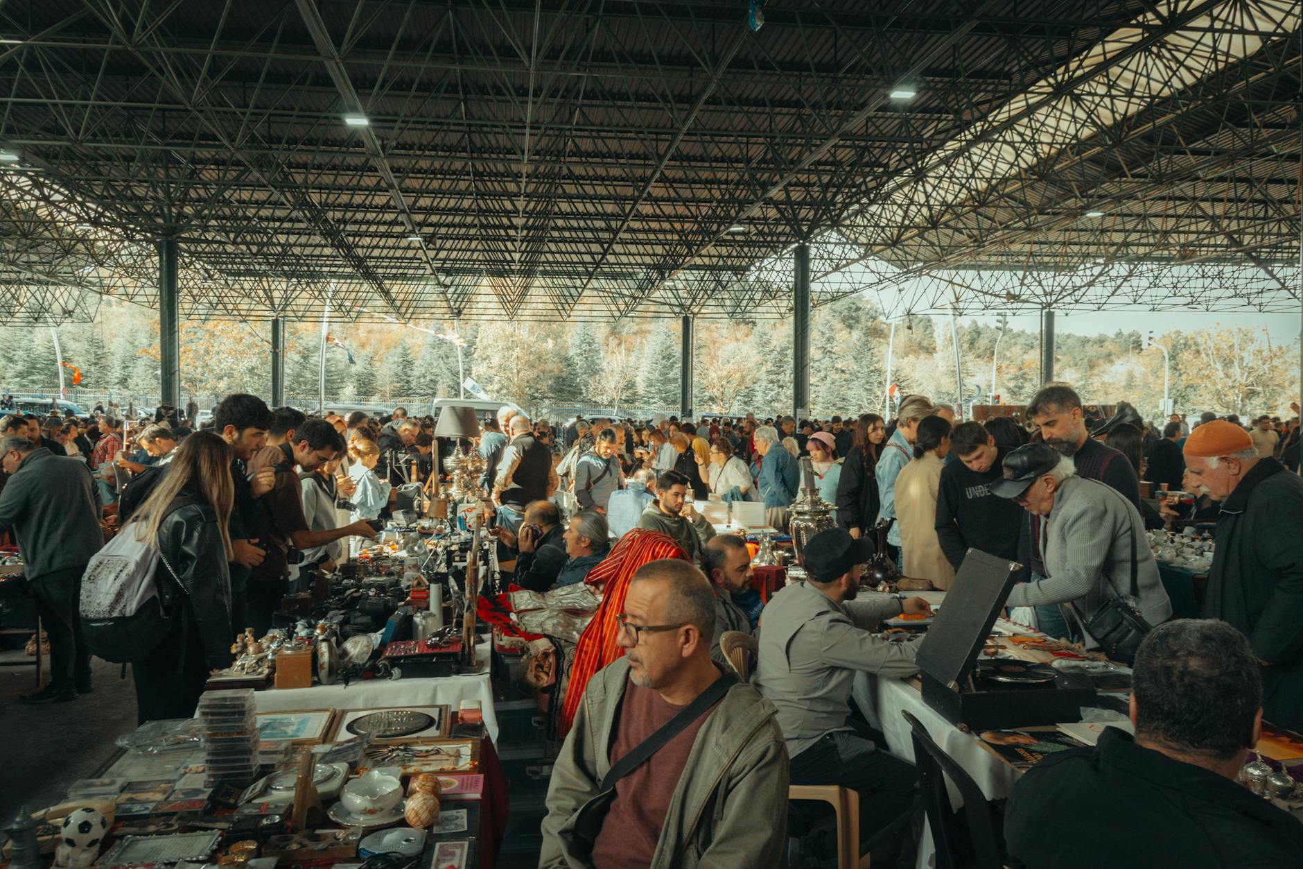 Bustling flea market with diverse crowd under a metal canopy, showcasing various goods.