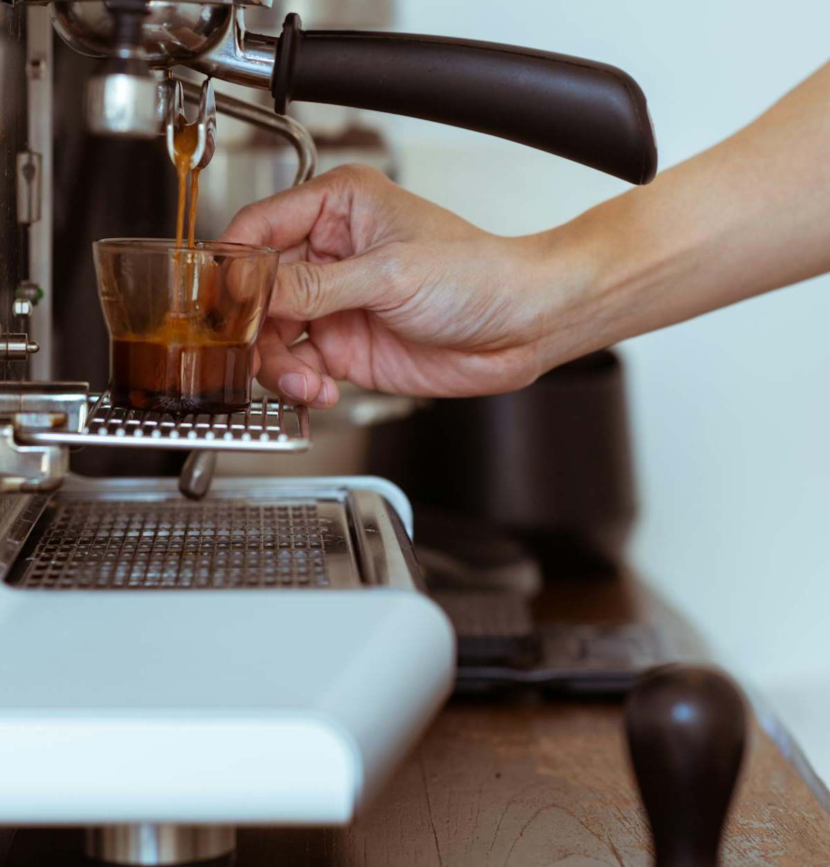 Close-up of a barista's hand pouring espresso from a machine into a glass cup.