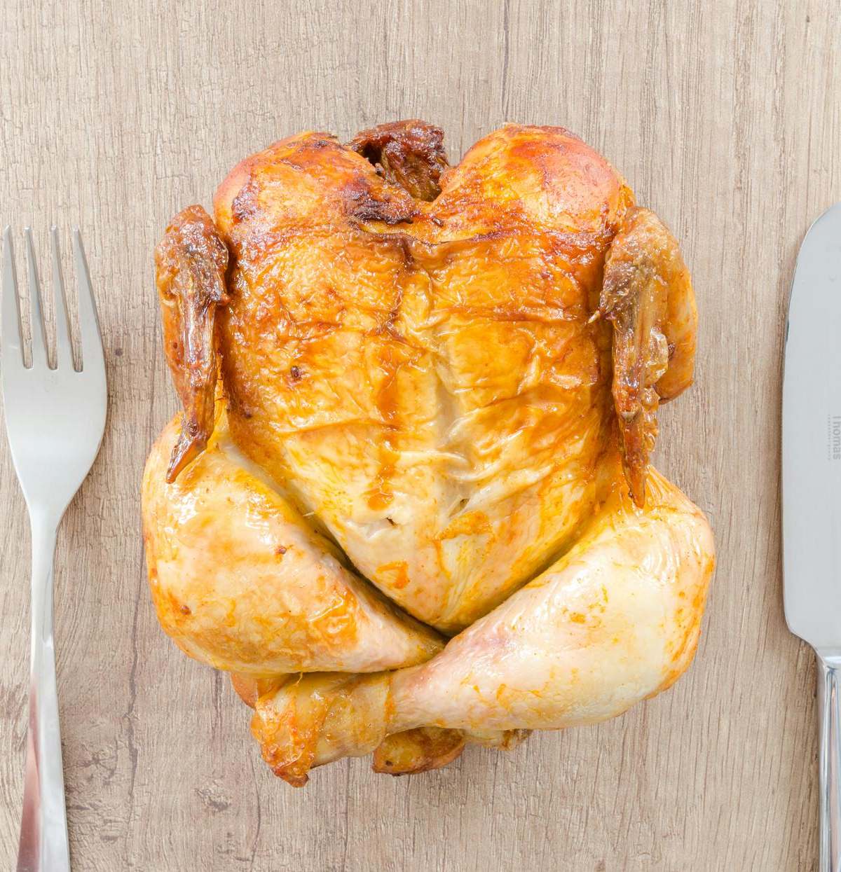 Top view of a perfectly roasted chicken placed between fork and knife on a wooden table.