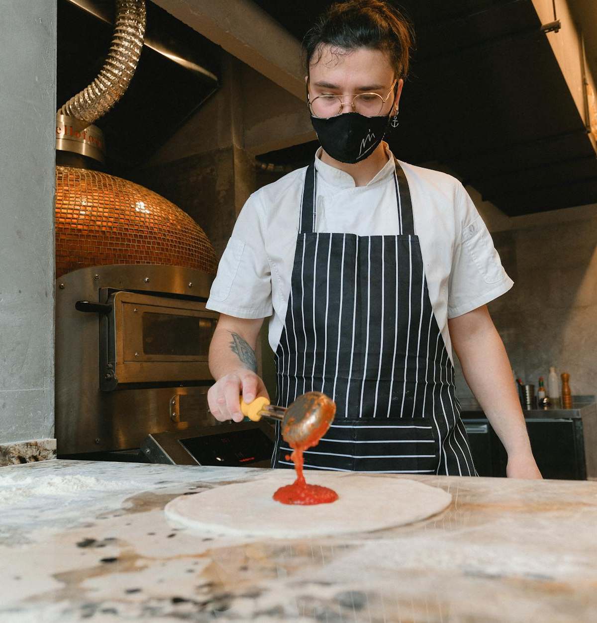 Chef wearing mask spreads tomato sauce on pizza base in kitchen.