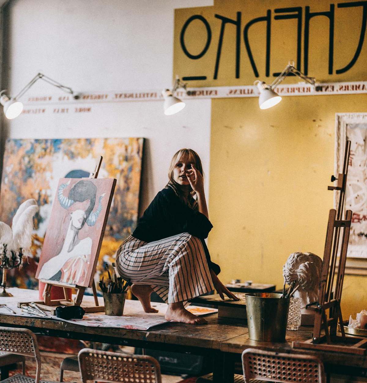 A woman artist sitting on a work table in a vibrant art studio surrounded by paintings and sculptures.