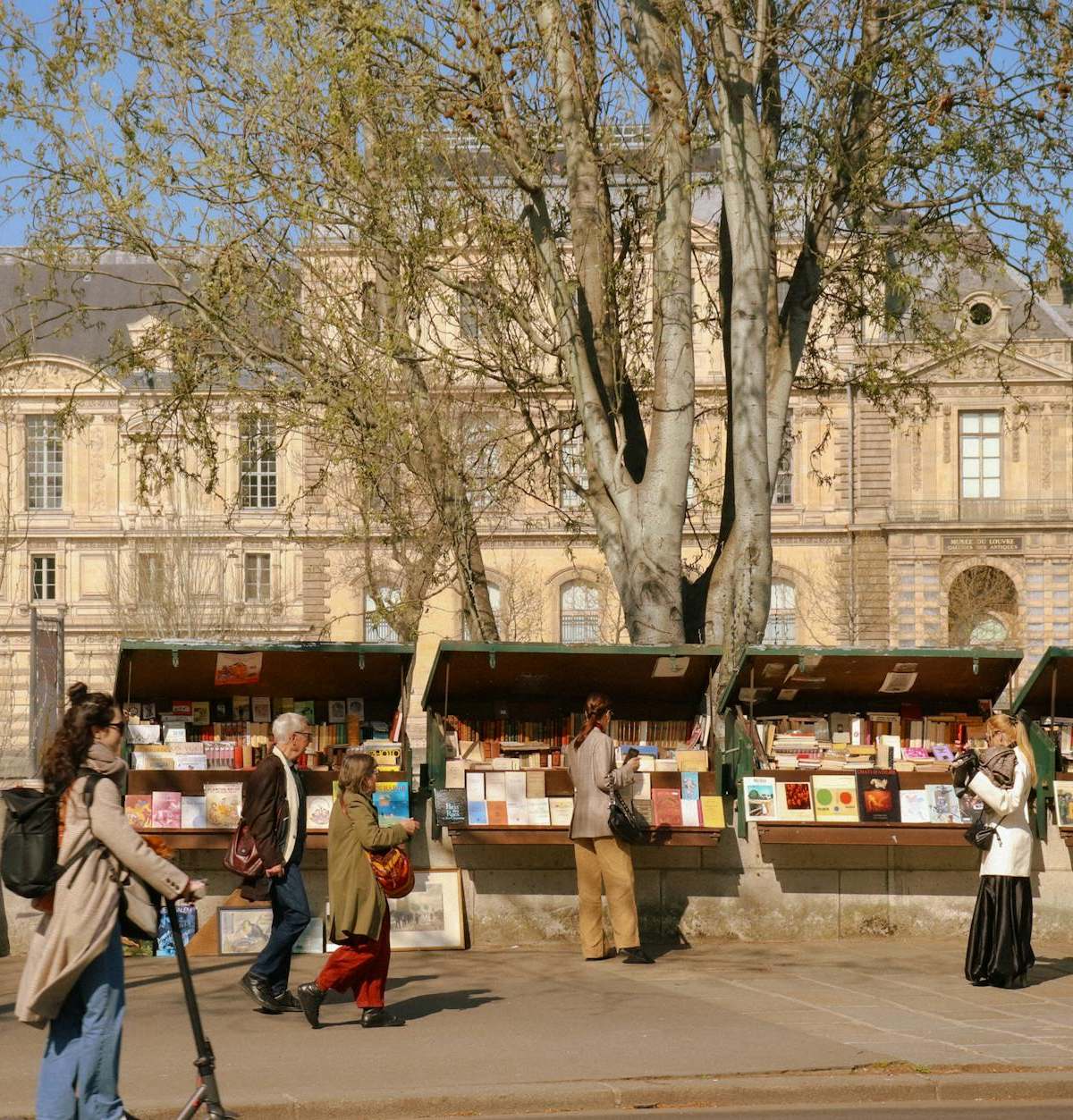 People browsing bookstalls by the Seine River in Paris on a sunny day.