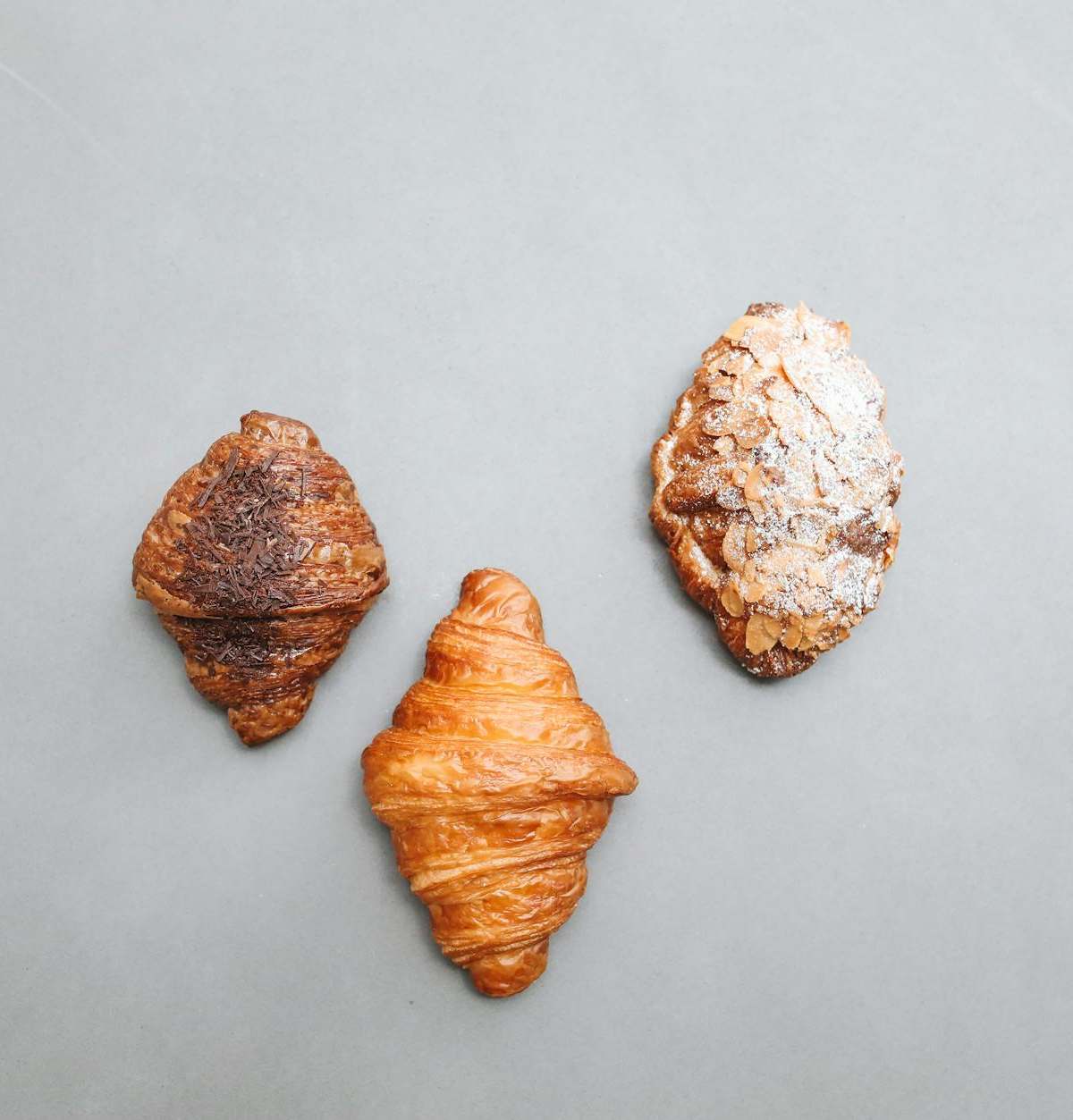 A trio of artisan croissants including plain, chocolate, and almond on a gray background in a flat lay style.