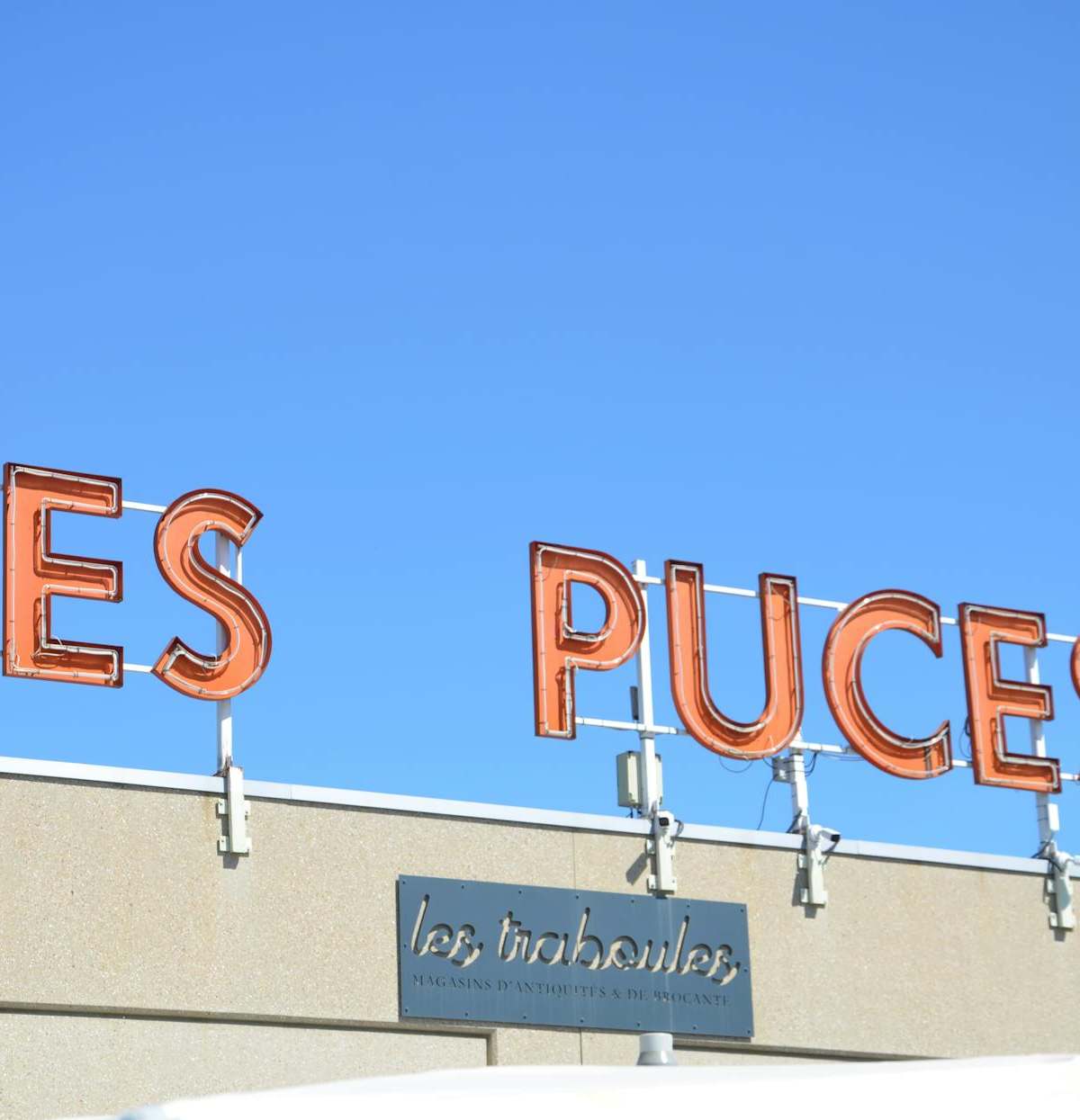 Bright neon 'Les Puces' sign on a building facade with a clear blue sky backdrop, France.