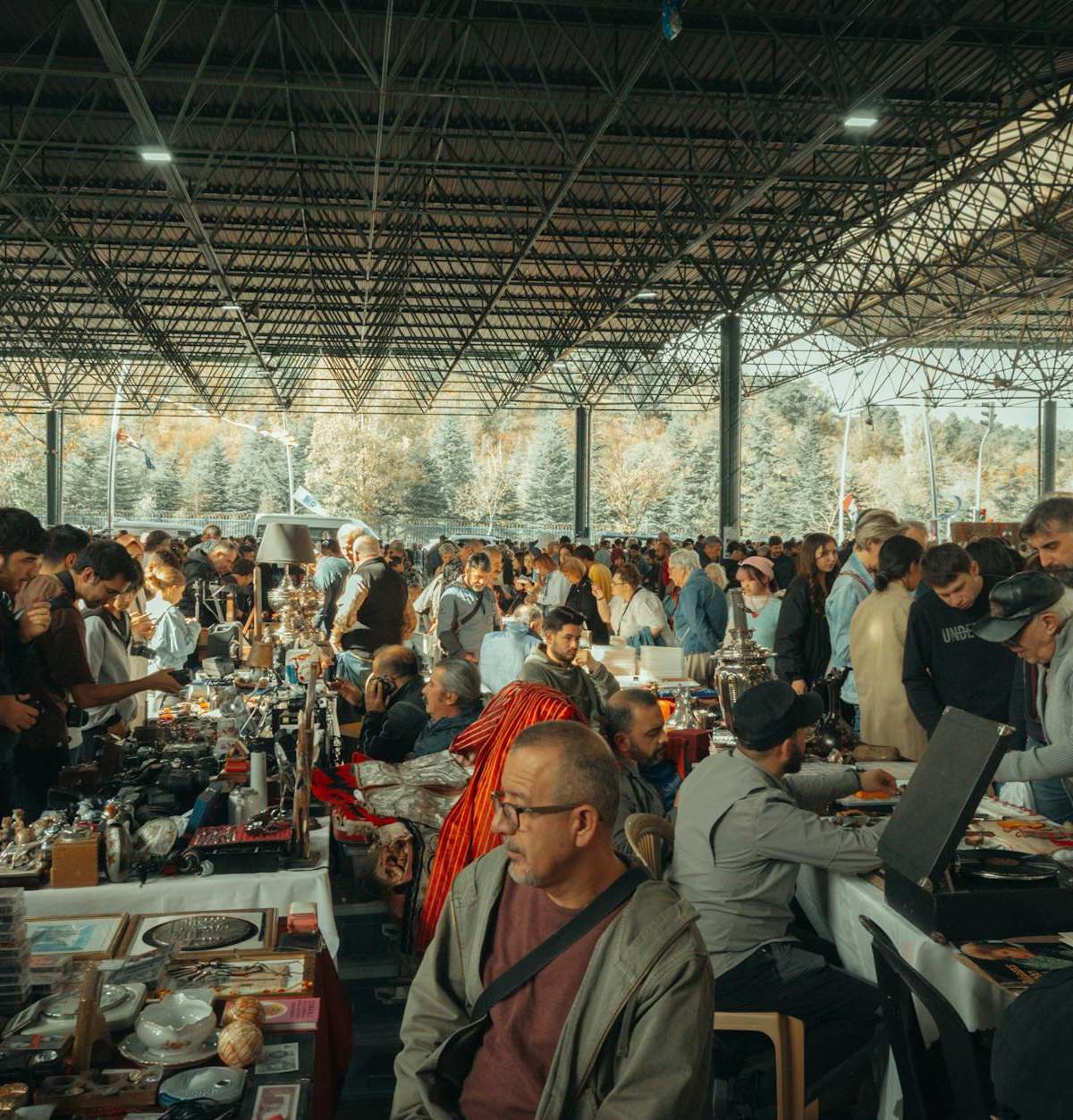 Bustling flea market with diverse crowd under a metal canopy, showcasing various goods.